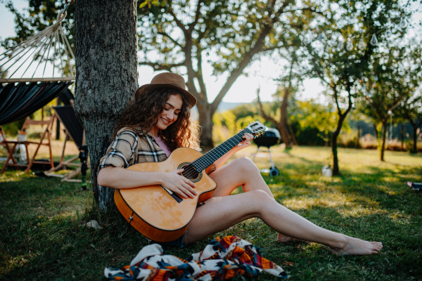 Beautiful woman playing guitar outside, sitting on the ground and leaning against tree. Solo camping trip in nature.