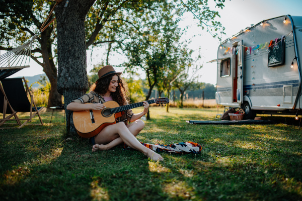 Young beautiful woman is on a camping trip in nature, sitting by big tree, playing guitar.