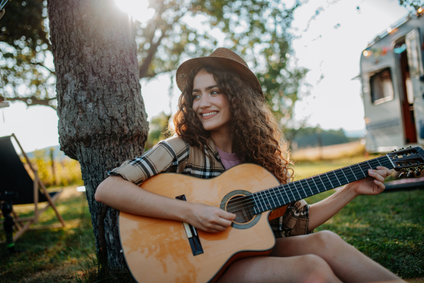 Young beautiful woman is on a camping trip in nature, sitting by tree, playing guitar