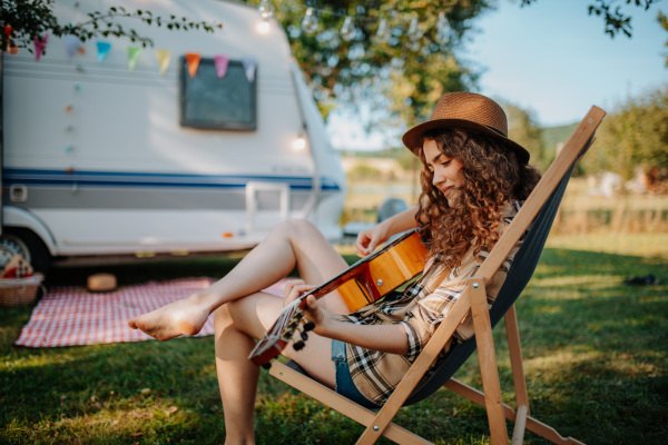 Young beautiful woman is on a camping trip in nature, sitting in front caravan, playing guitar.