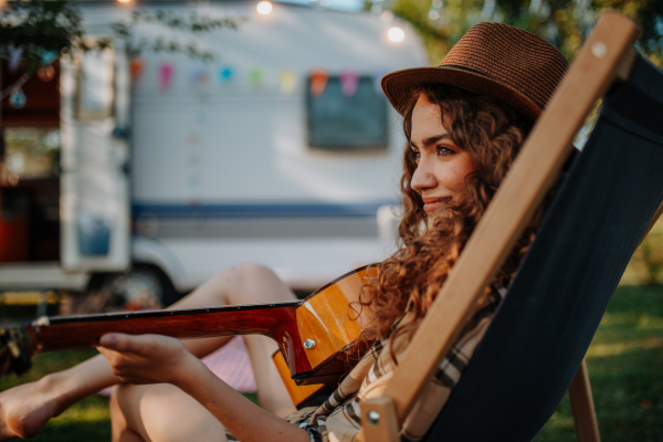 Young beautiful woman is on a camping trip in nature, sitting in front caravan, playing guitar.