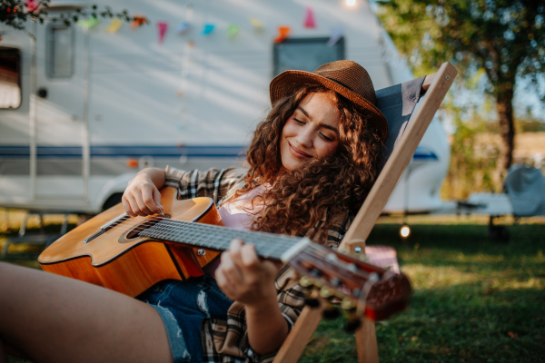 Young beautiful woman is on a camping trip in nature, sitting in front caravan, playing guitar.