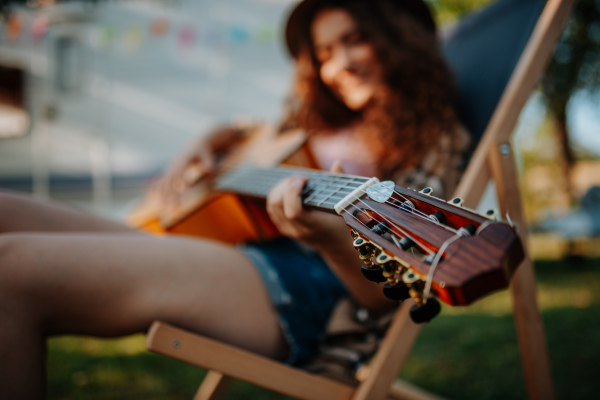 Young beautiful woman is on a camping trip in nature, sitting in front caravan, playing guitar.