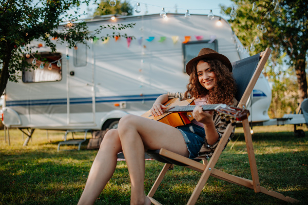 Young beautiful woman is on a camping trip in nature, sitting in front caravan, playing guitar.