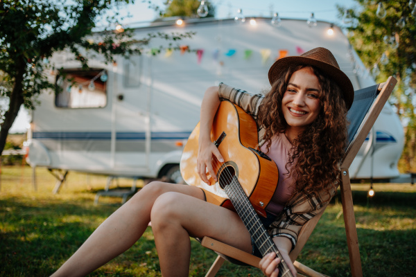 Young beautiful woman is on a camping trip in nature, sitting in front caravan, playing guitar.