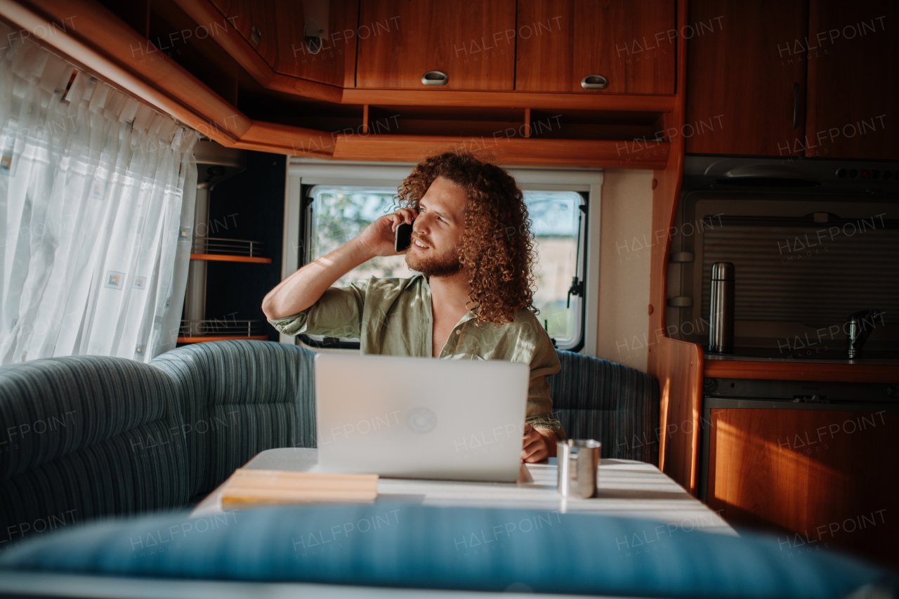 Young man working remotely from caravan, making phone call. Man using camper as homeoffice, working on notebook