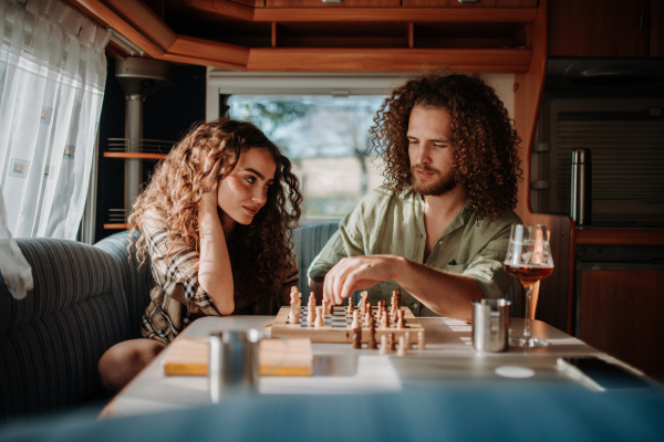 Young couple is on a camping trip in nature, sitting in caravan and playing chess.