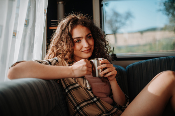 Young beautiful woman is on a camping trip in nature, sitting in her caravan and enjoying morning cup of coffee