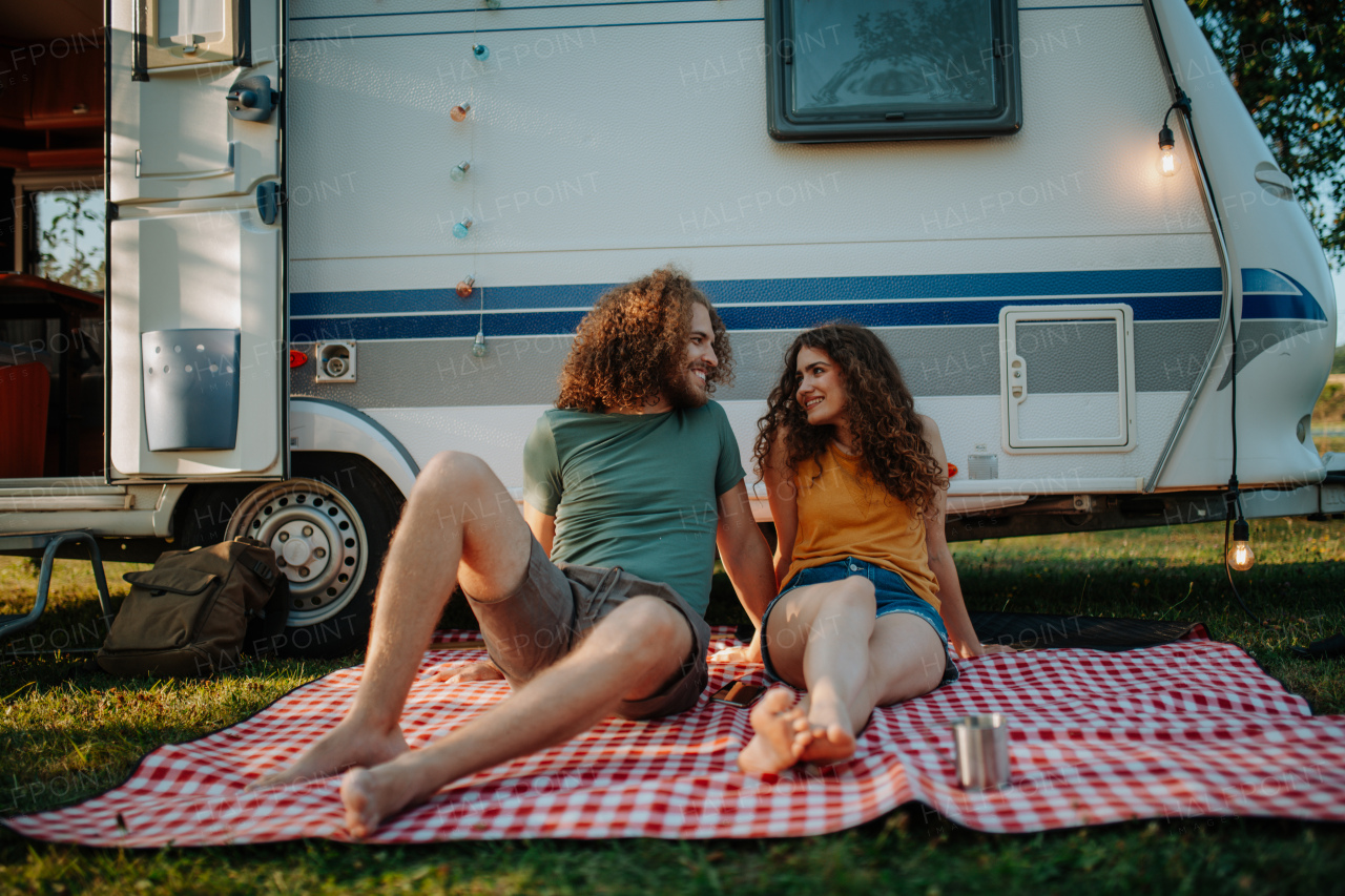 Beautiful couple sitting in front of caravan looking at each other lovingly, enjoying peaceful moment. Camping trip for young people.