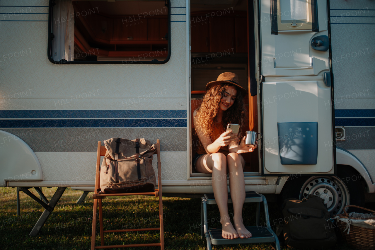 Portrait of young beautiful woman on a camping trip in nature, sitting on caravan steps with smartphone and coffee in her hand.