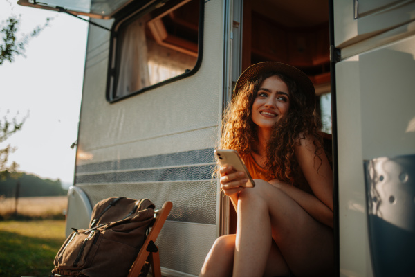 Portrait of young beautiful woman on a camping trip in nature, sitting on caravan steps with smartphone in her hand.