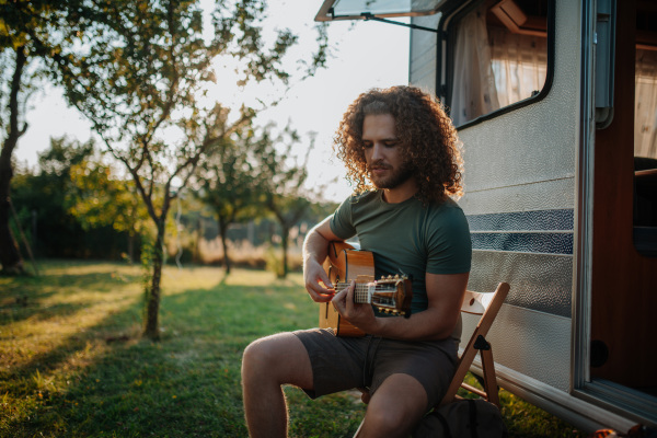 Young man is on a camping trip in nature alone, sitting in front caravan and playing guitar