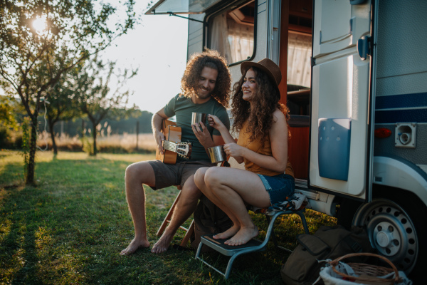 Couple sitting by caravan, man playing guitar and woman working on laptop.