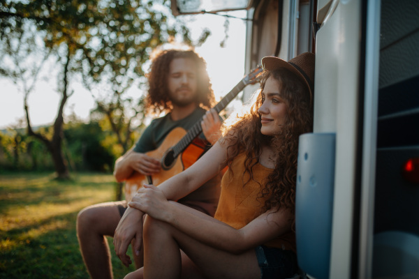 Couple sitting on caravan steps, enjoying peaceful moment. Man playing guitar for his girlfriend.