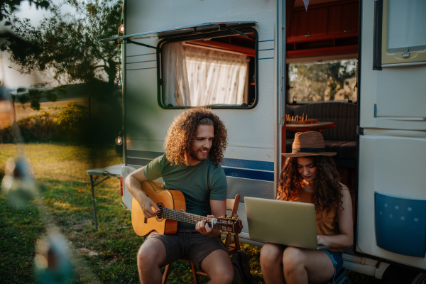 Couple sitting by caravan, man playing guitar and woman working on laptop.