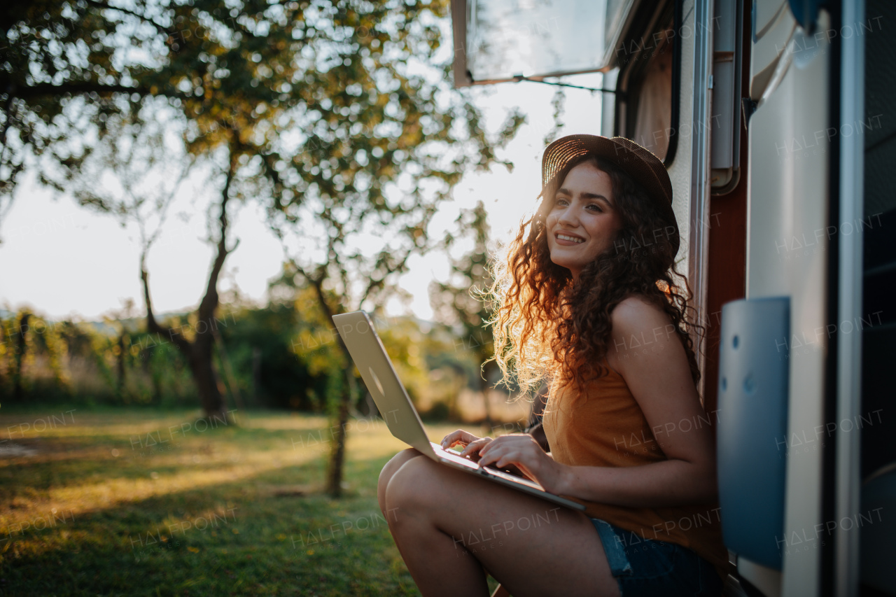 Young beautiful woman is on a camping trip in nature, sitting in front of caravan with notebook on knees. Caravan as homeoffice, working remotely