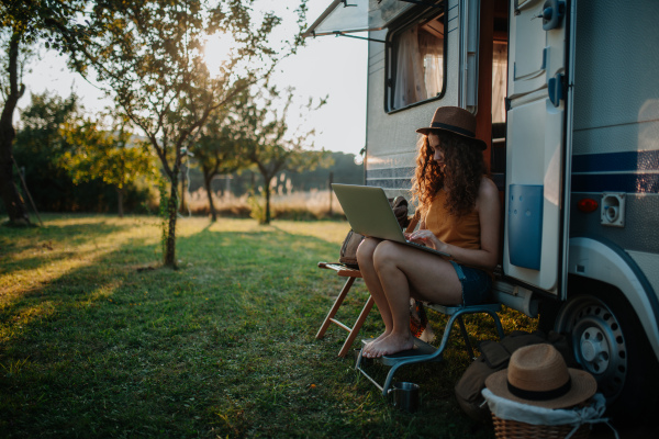 Young beautiful woman is on a camping trip in nature, sitting in front of caravan with notebook on knees. Caravan as homeoffice, working remotely