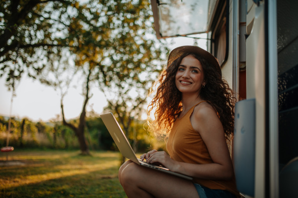 Young beautiful woman is on a camping trip in nature, sitting in front of caravan with notebook on knees. Caravan as homeoffice, working remotely