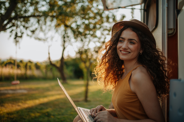 Young beautiful woman is on a camping trip in nature, sitting in front of caravan with notebook on knees. Caravan as homeoffice, working remotely