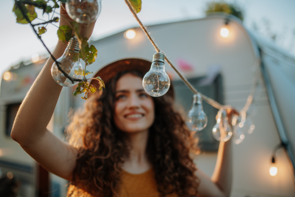 Young beautiful woman is on a camping trip in nature, putting lights on tree, preparing for evening