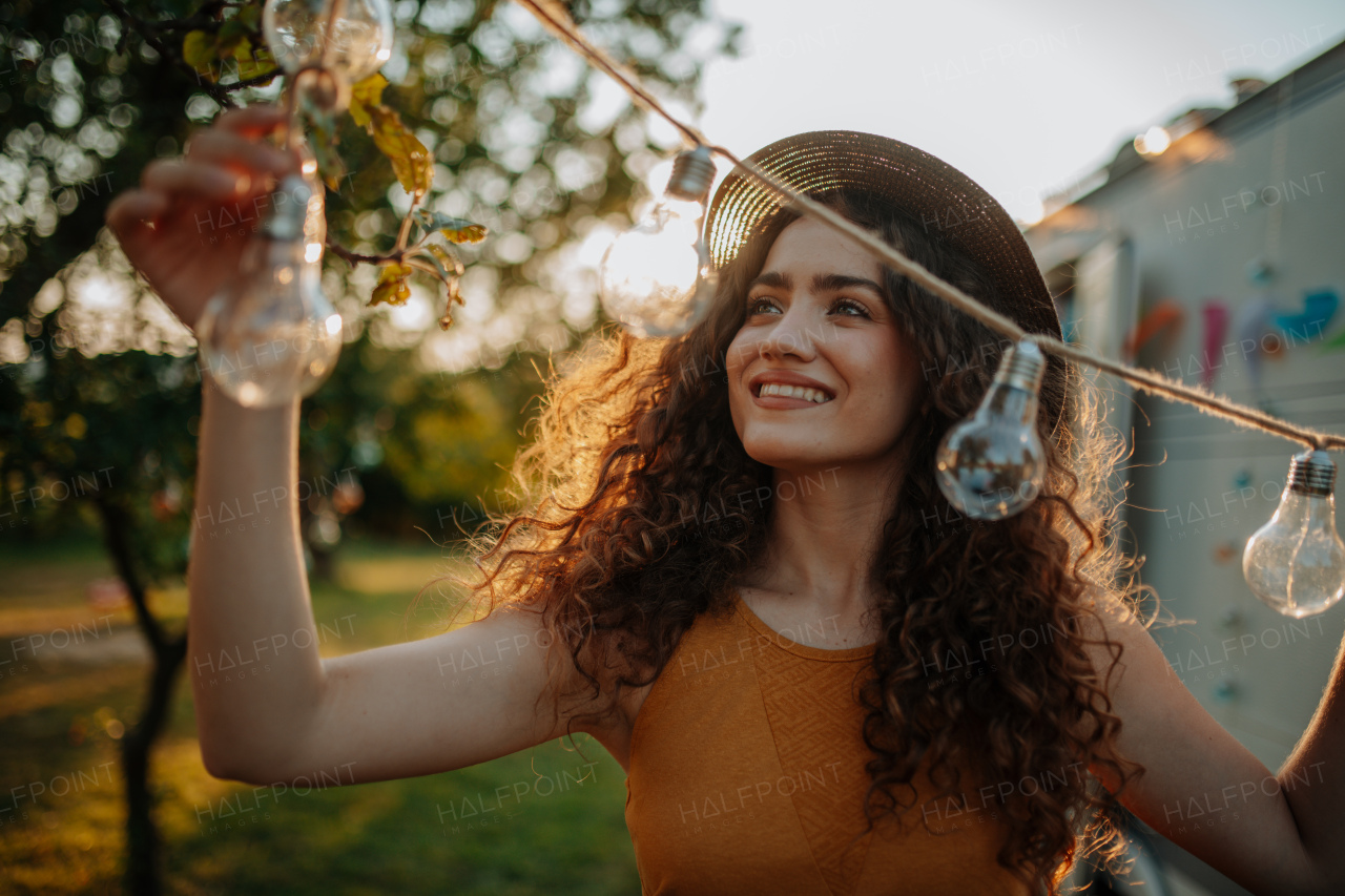 Young beautiful woman is on a camping trip in nature, putting lights on tree, preparing for evening