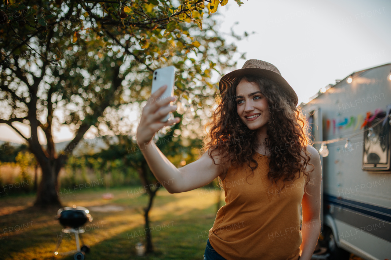 Portrait of young beautiful woman on camping trip in nature, standing in front of caravan, holding smartphone, , taking selfie photo