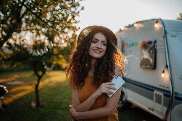 Portrait of young beautiful woman on camping trip in nature, standing in front of caravan, holding smartphone