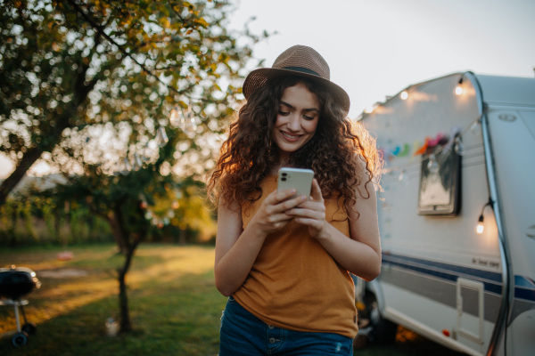 Portrait of young beautiful woman on camping trip in nature, standing in front of caravan, holding smartphone