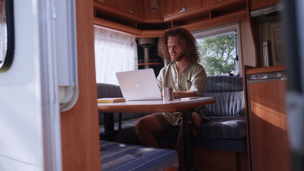 Young man sitting inside of a caravan and working on his laptop. Concept of remote work.