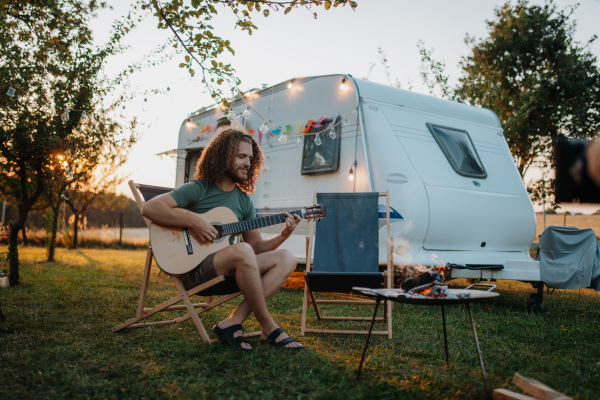 Young man is on a camping trip in nature alone, sitting in front caravan and playing guitar