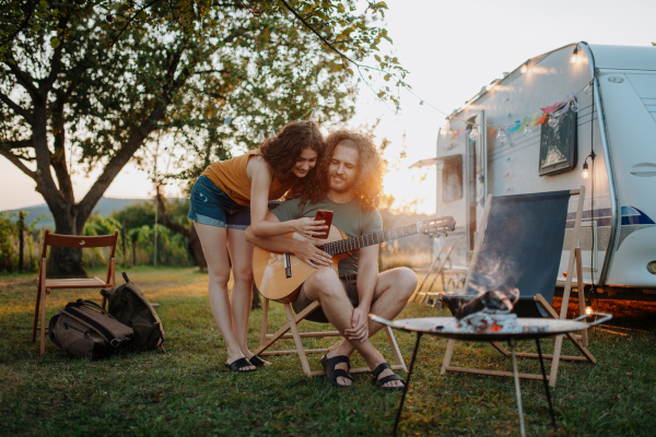 Couple sitting by campfire in the evening, enjoying peaceful moment. Man playing guitar and singing song to his girlfriend.