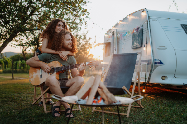 Couple sitting by campfire in the evening, enjoying peaceful moment. Man playing guitar and singing song to his girlfriend.