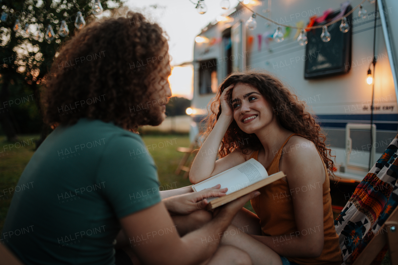 Couple having romantic camping trip in nature. Man reading book to girlfriend. Caravan traveling for young people