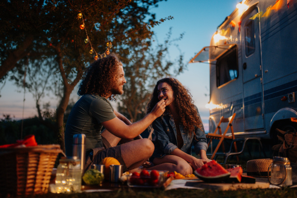 Couple having romantic picnic during camping trip in nature. Man feeding his girlfriend. Caravan traveling for young people