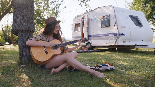 Young man on a roadtrip with a caravan, camping and exploring new places. Sitting in front of caravan and playing guitar.