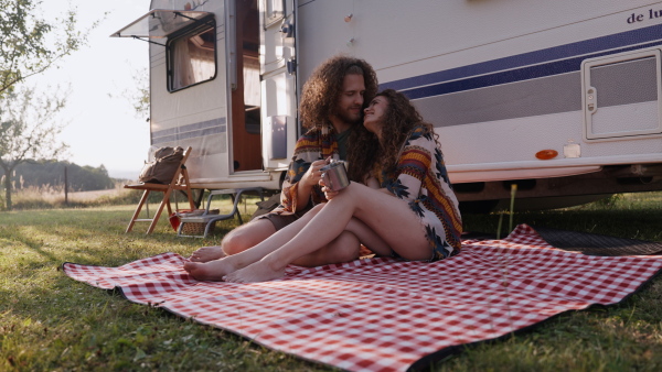 Young couple in love on a roadtrip with a caravan, camping and exploring new places. Sitting on blanket, drinking tea.