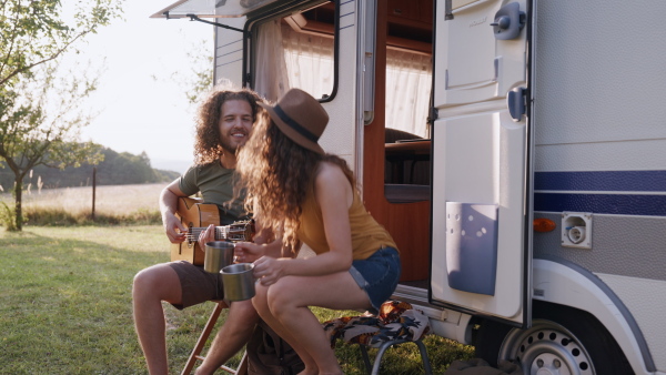 Young couple in love on a roadtrip with a caravan, camping and exploring new places. Sitting in front of caravan and playing guitar.