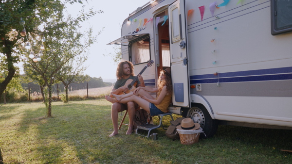 Young couple in love on a roadtrip with a caravan, camping and exploring new places. Sitting in front of caravan and playing guitar.