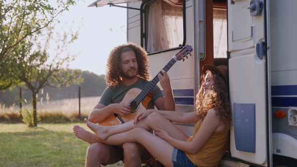 Young couple in love on a roadtrip with a caravan, camping and exploring new places. Sitting in front of caravan and playing guitar.