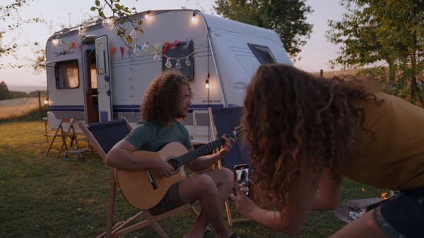 Young couple in love on a roadtrip with a caravan, camping and exploring new places. Sitting in front of caravan and playing guitar while girlfirend making a video.