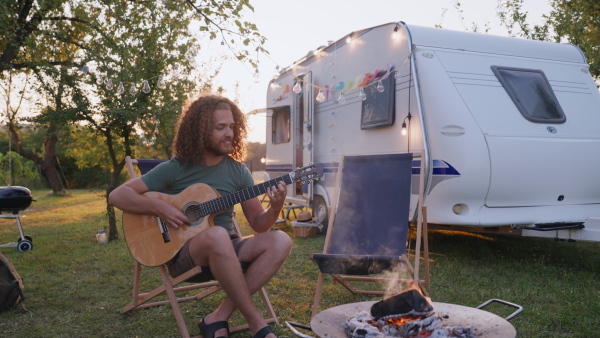 Young man on a roadtrip with a caravan, camping and exploring new places. Sitting in front of caravan and playing guitar.