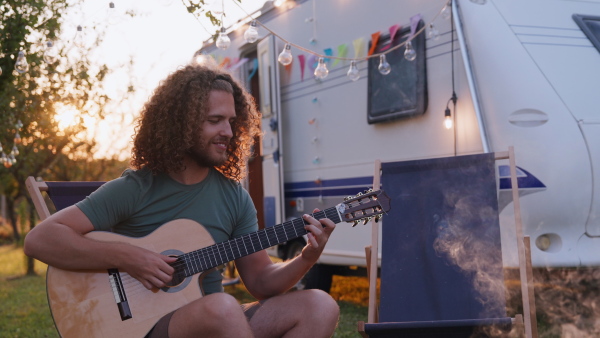 Young man on a roadtrip with a caravan, camping and exploring new places. Sitting in front of caravan and playing guitar.