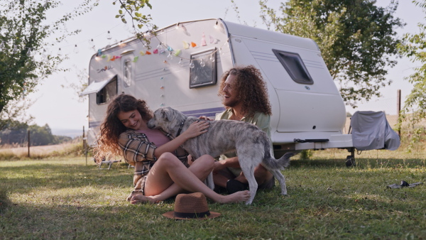 Young couple in love on a roadtrip with a caravan, camping and exploring new places. Sitting in front of caravan and playing with dog.