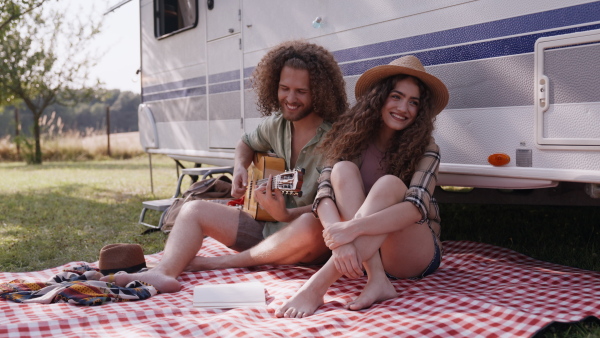 Young couple in love on a roadtrip with a caravan, camping and exploring new places. Sitting in front of caravan and playing guitar.