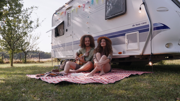 Young couple in love on a roadtrip with a caravan, camping and exploring new places. Sitting in front of caravan and playing guitar.