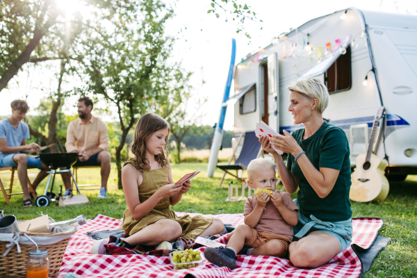 Family having picnic in front of caravan, sitting on picnic blanket and grilling diner on outdoor grill. Family trip in caravan