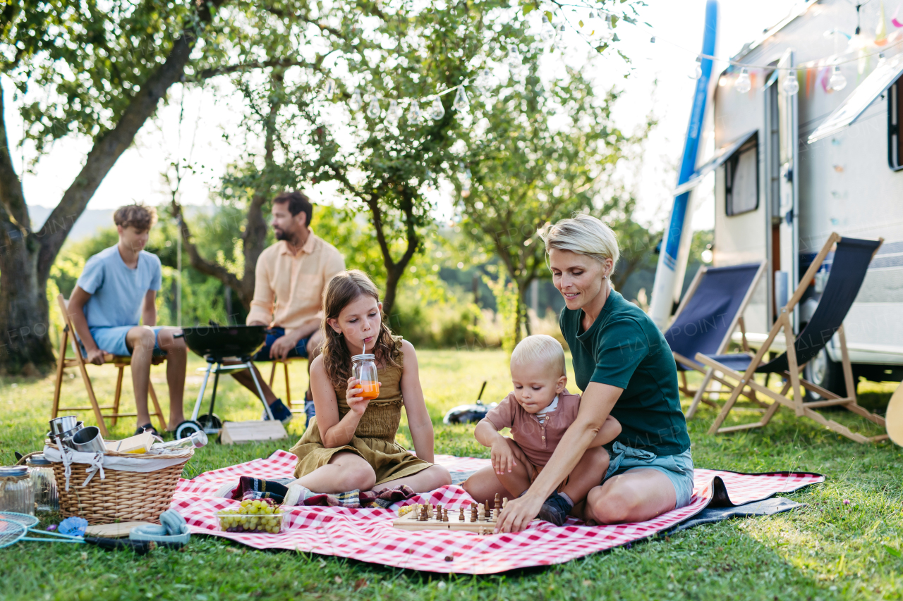 Family having picnic in front of caravan, sitting on picnic blanket and grilling diner on outdoor grill. Family trip in caravan