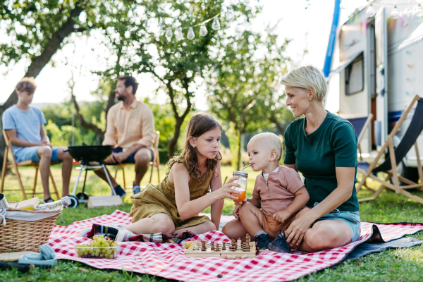 Family having picnic in front of caravan, sitting on picnic blanket and grilling diner on outdoor grill. Family trip in caravan