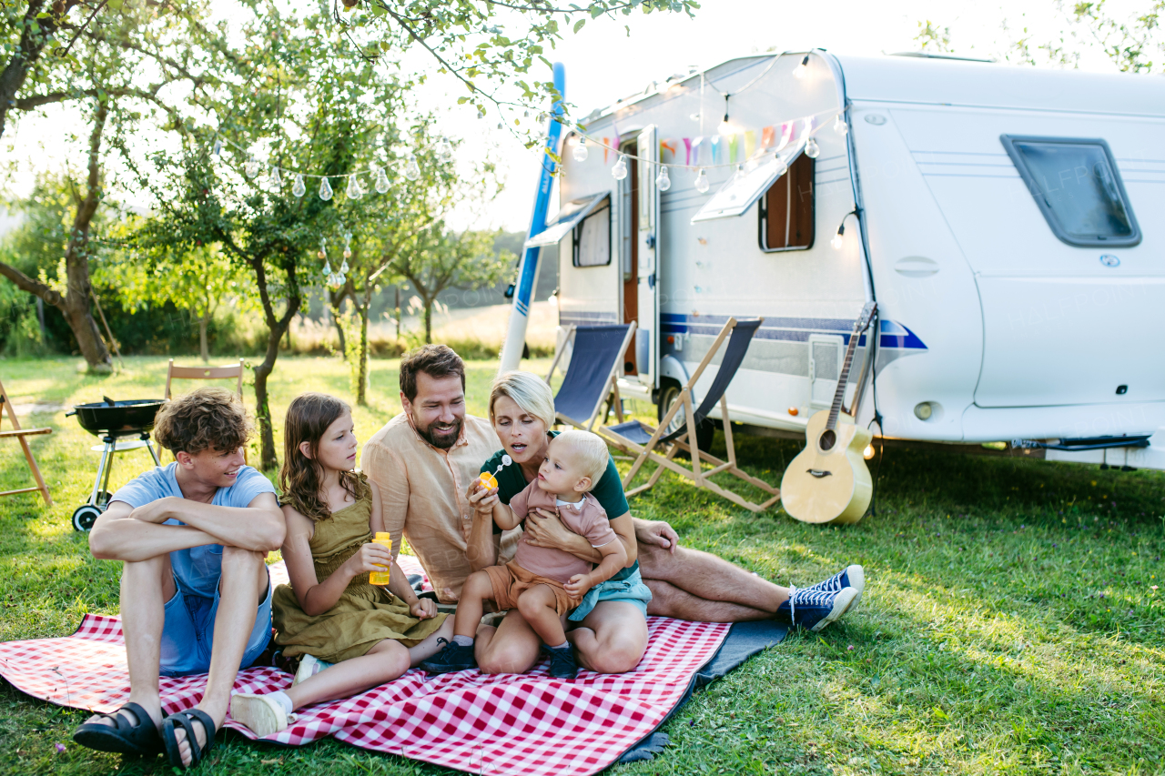 Family having picnic in front of caravan, sitting on picnic blanket and blowing bubbles. Family trip in caravan