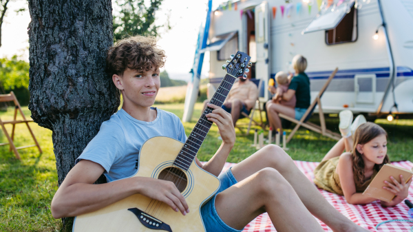 Family spending time outdoor, camping together. Teenage boy playing guitar in front of caravan.